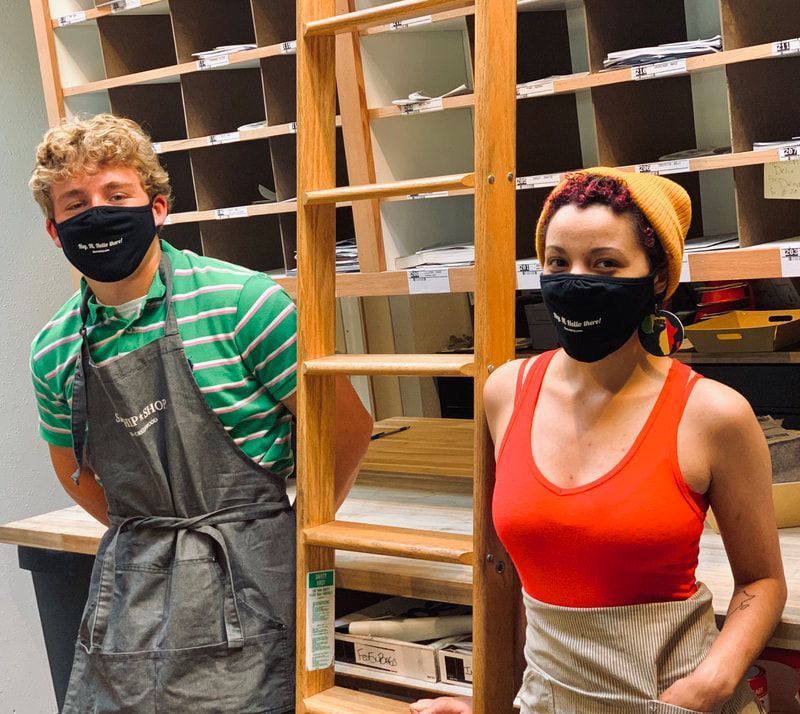 Two masked people in aprons stand in front of empty shelves in a storage room.