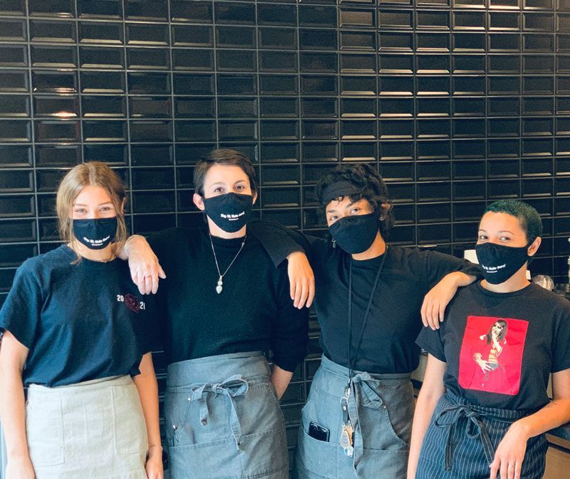 Four masked friends standing arm-in-arm in front of a dark brick wall.