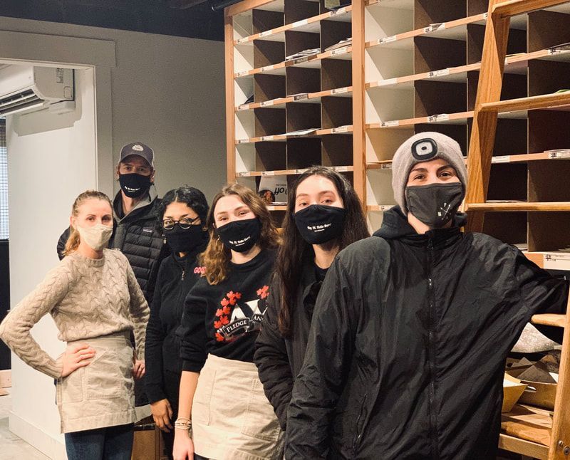 Five masked people stand in a warehouse-like room with shelves of cardboard bins and an air conditioner.