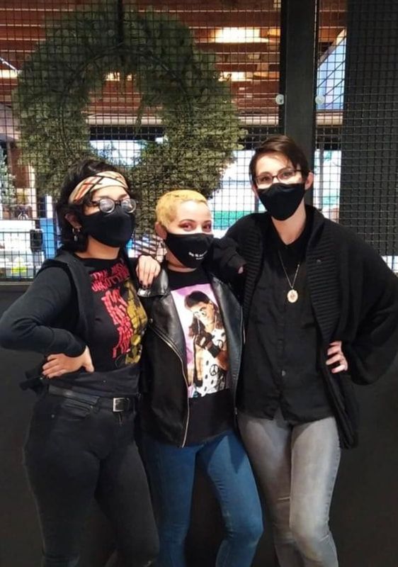 Three masked people posing together indoors, with a large green wreath behind them.
