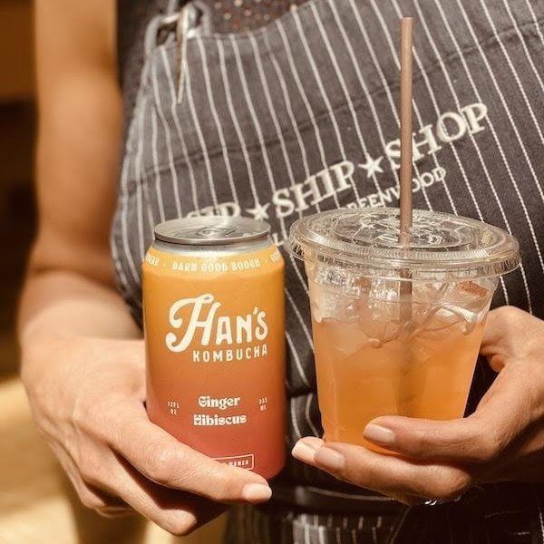 Hands holding a Hans Kombucha can and a clear iced drink with a straw and bamboo spoon
