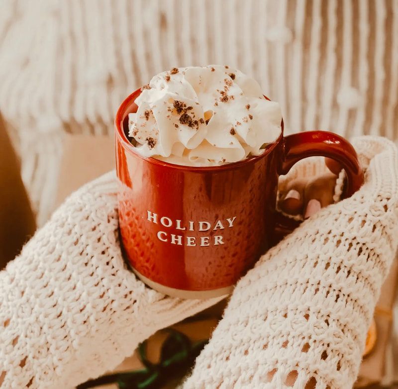 Hands holding a red mug of whipped cocoa labeled “Holiday Cheer” on a wooden table