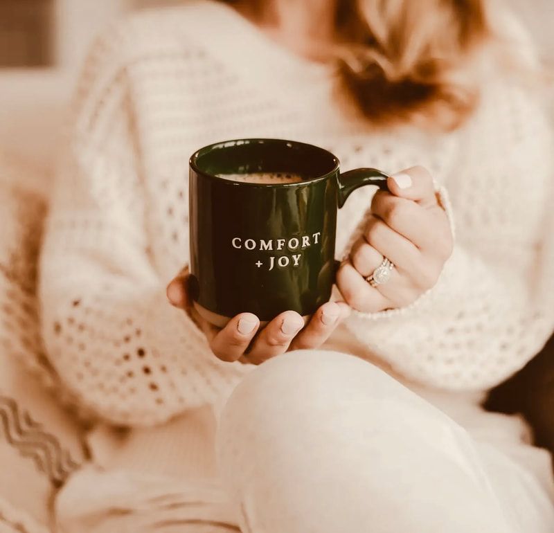 Person in a cream sweater holding a dark green mug that says “COMFORT + JOY”