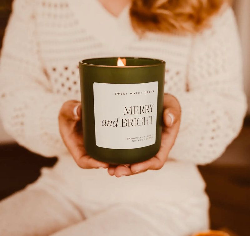 Person holding a lit green candle labeled “Merry and Bright” in a cozy knit sweater