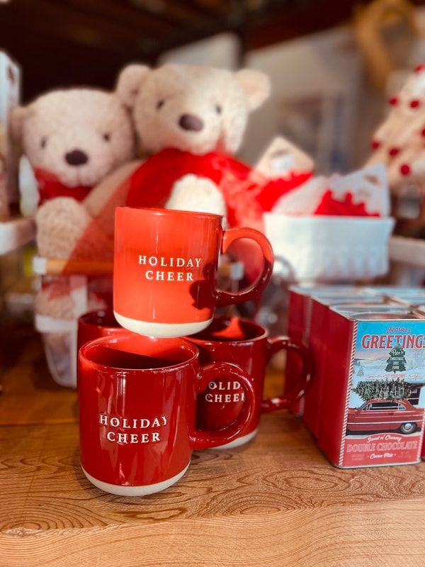 Red holiday mugs on a table with teddy bears, tissue paper, and a small product box behind them