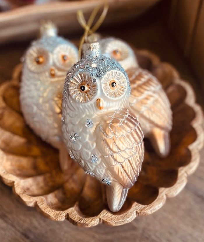 Two small owl figurines in a carved wooden dish, white and gold with large round eyes.