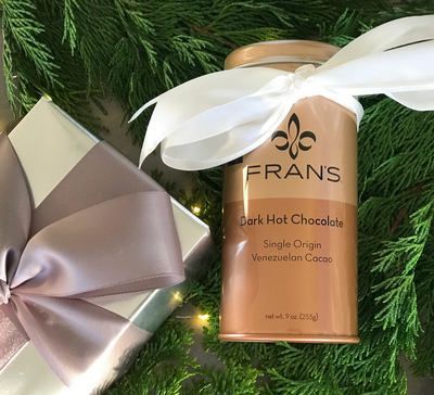 Gold FRANS hot chocolate tin with white ribbon gift box on evergreen branches and lights