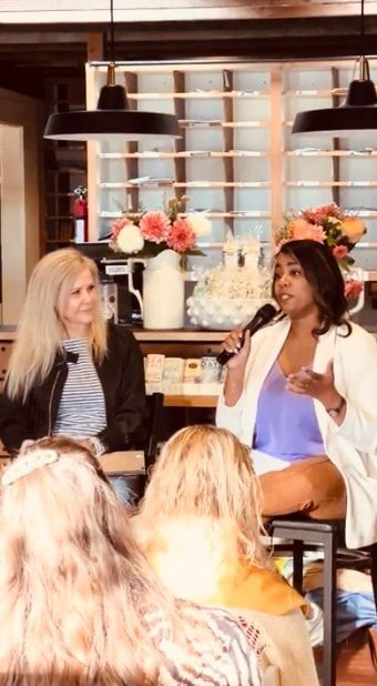 Two women speaking at a panel event, seated before a floral backdrop and audience.
