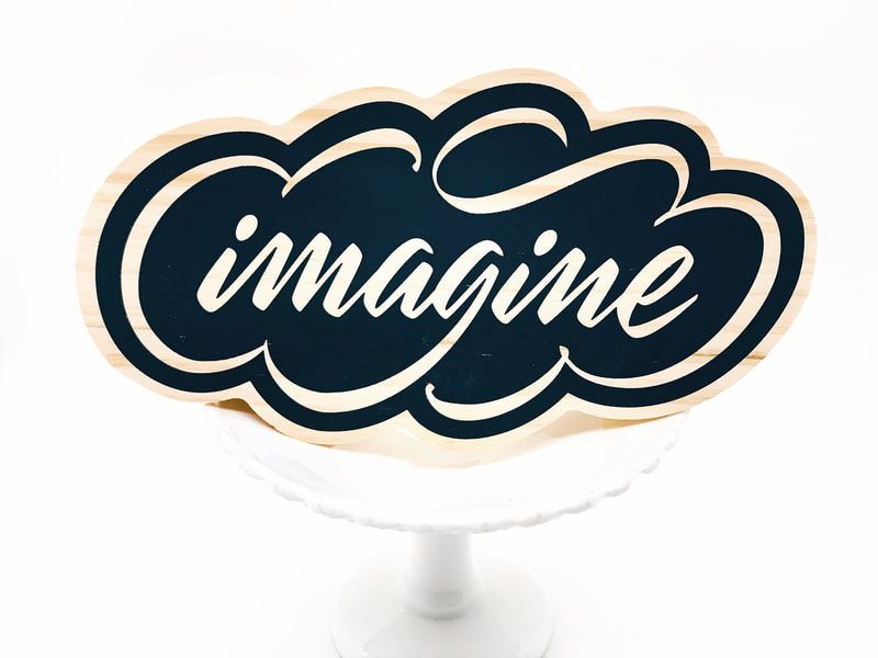 Decorative sign with the word “imagine” in white script on a dark navy cloud shape with a cream border