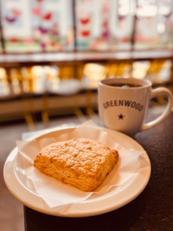 A pastry on a white plate beside a steaming GREENWOOD coffee mug in a cozy cafe.