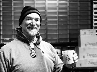 Smiling person in a beanie holding a mug in front of a shuttered storefront