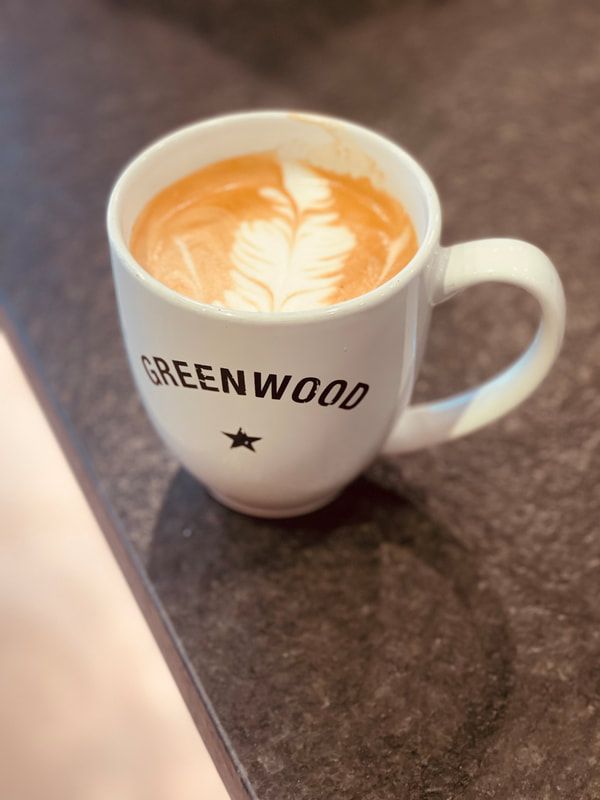 White mug of latte with leaf latte art on a dark countertop, labeled “GREENWOOD.”
