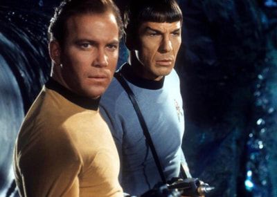 Two Star Trek crew members in yellow and blue uniforms stand against a dark starship backdrop.