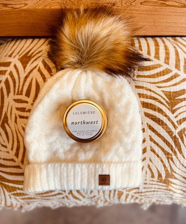 White knit beanie with faux fur pom-pom and logo patch, laid on a patterned blanket