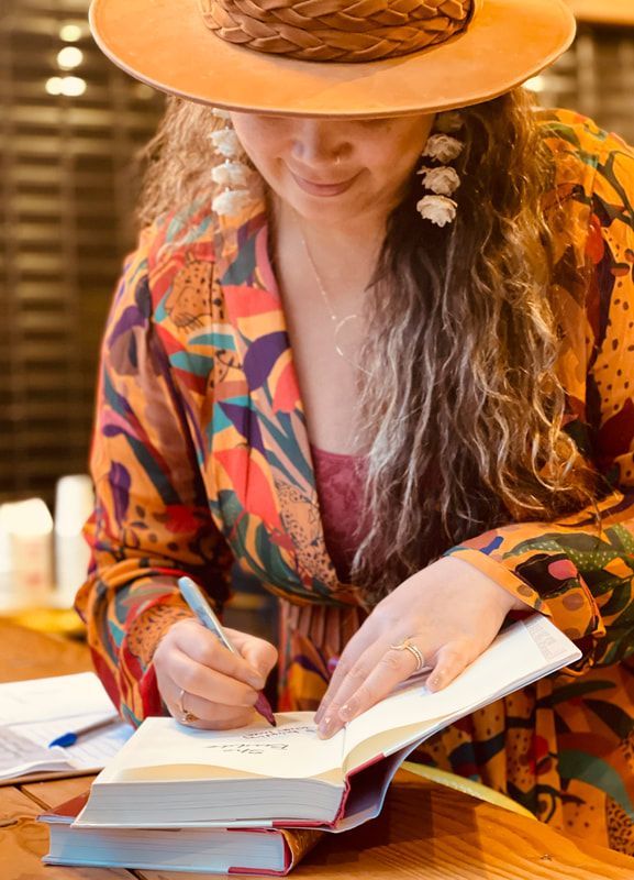 Person in a colorful hat and patterned shirt writing in an open notebook at a table.