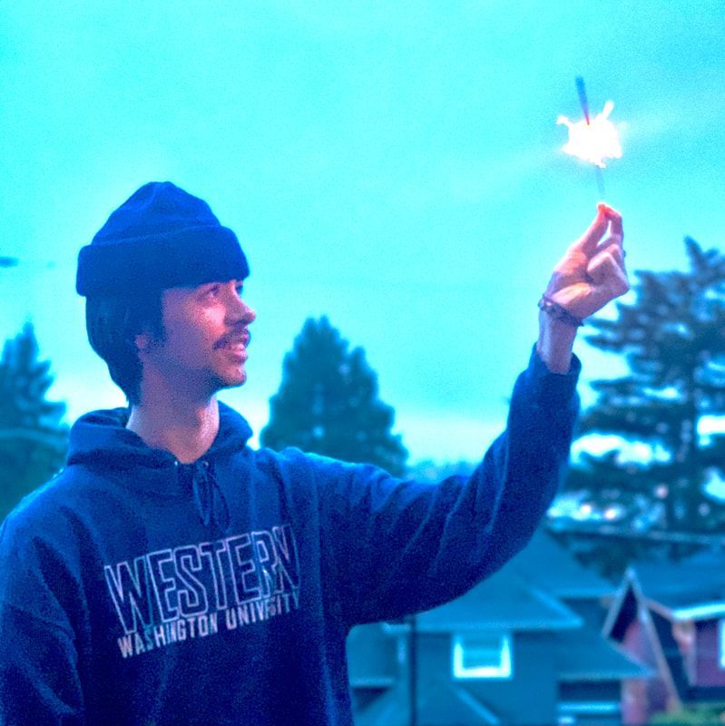 Person in a blue hoodie holds up a lit sparkler outdoors at dusk.