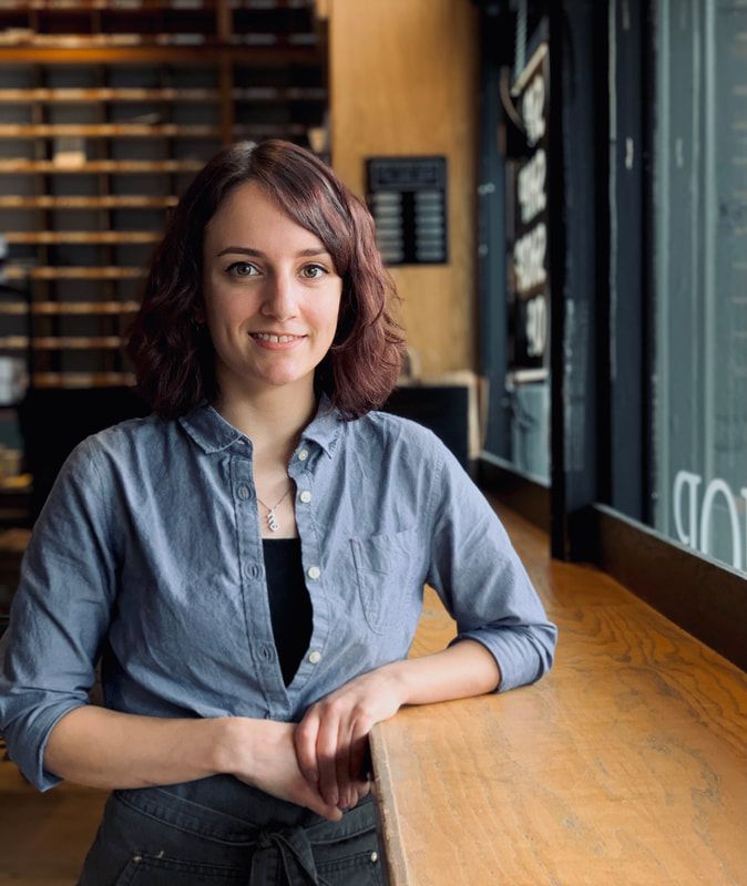 Person sitting at a wooden table in a cafe, smiling at the camera with windows and shelves behind them.