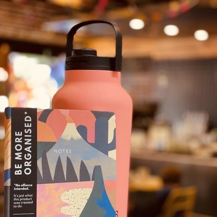 Coral water bottle beside a notebook with a bold “Be More Organised” cover in a blurred cafe setting