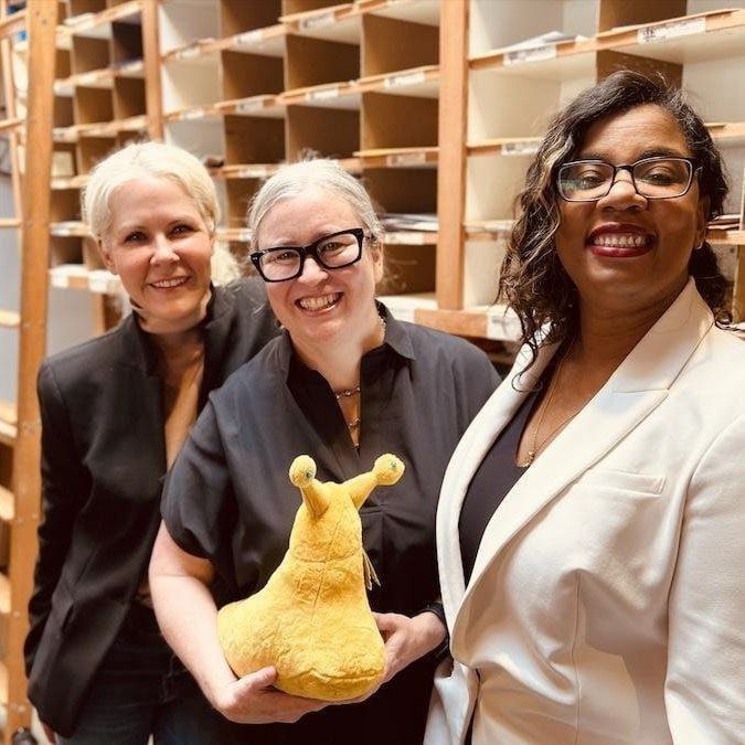Three smiling people in a library, holding a yellow plush toy in front of shelves