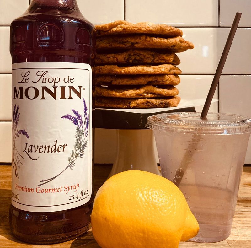 Lavender syrup bottle beside cookies, lemon, and iced drink with straw on a counter