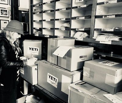 Worker packing YETI boxes in a warehouse aisle lined with shelves and stacked packages