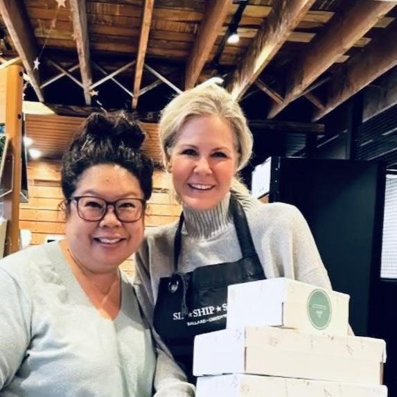 Two women smiling in a rustic kitchen, standing beside stacked white boxes in aprons.