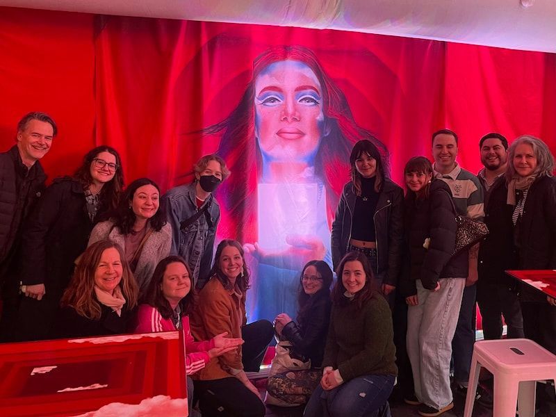 Group posing in front of a red-lit mural with a large illuminated face, smiling indoors