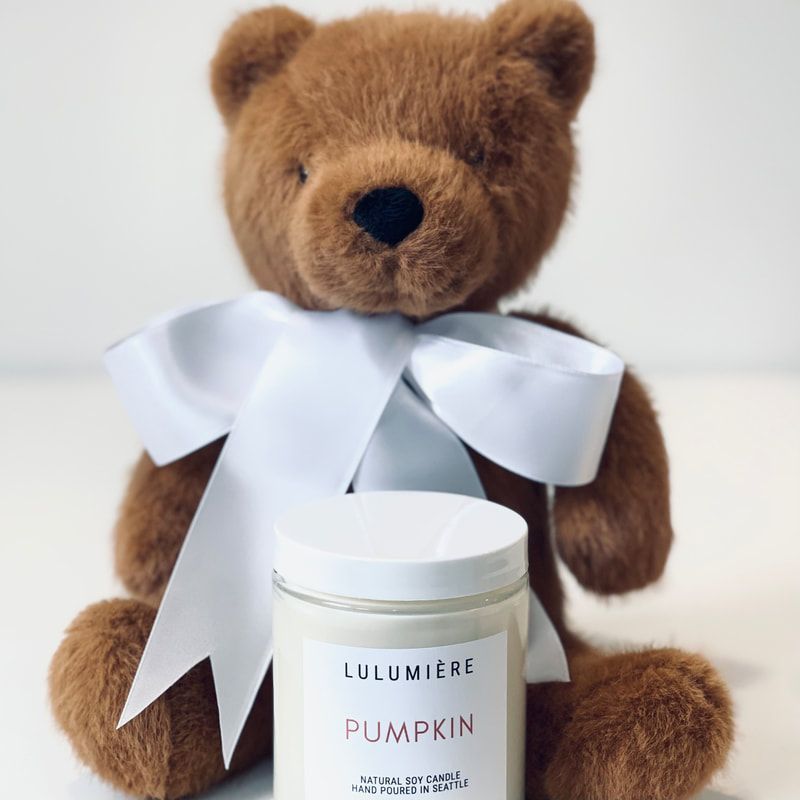 Brown teddy bear with a white ribbon sitting behind a white pumpkin-scented candle jar
