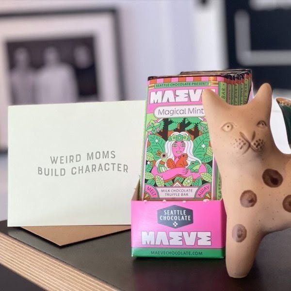 Product display with a Maeve comic box, pink case, tan figurine, and a card reading “Weird moms build character”