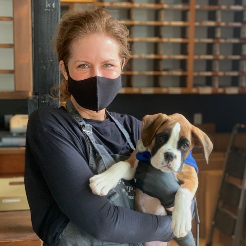 Person holding a brown-and-white puppy indoors, both wearing black masks.