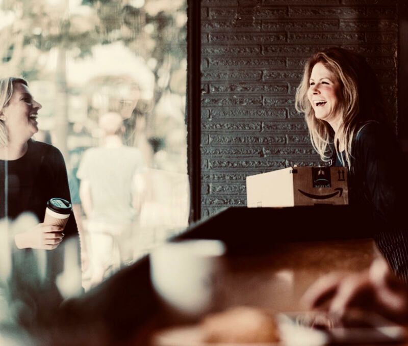 Two people chatting in a cafe, one holding a coffee cup and the other smiling by a brick wall.