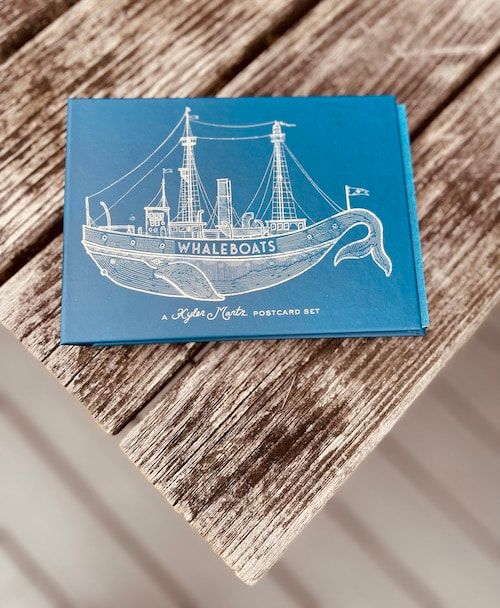 Blue Whaleboats card with a white line drawing of a ship on a wooden table