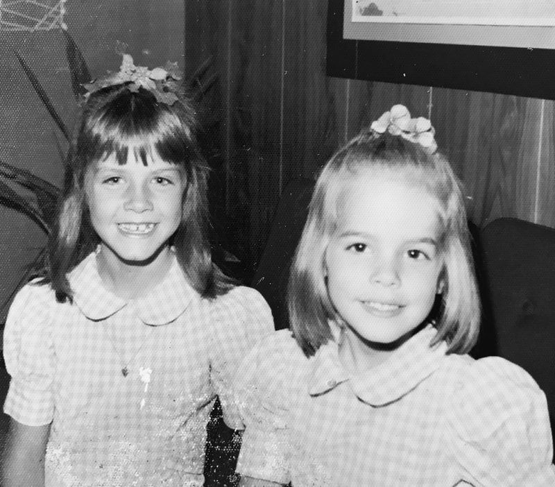 Two smiling children pose indoors in plaid dresses, standing close together.