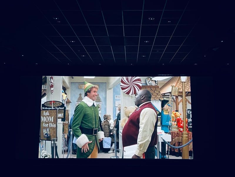 Two people in festive holiday attire chatting in a decorated shop with candy canes and seasonal decor.