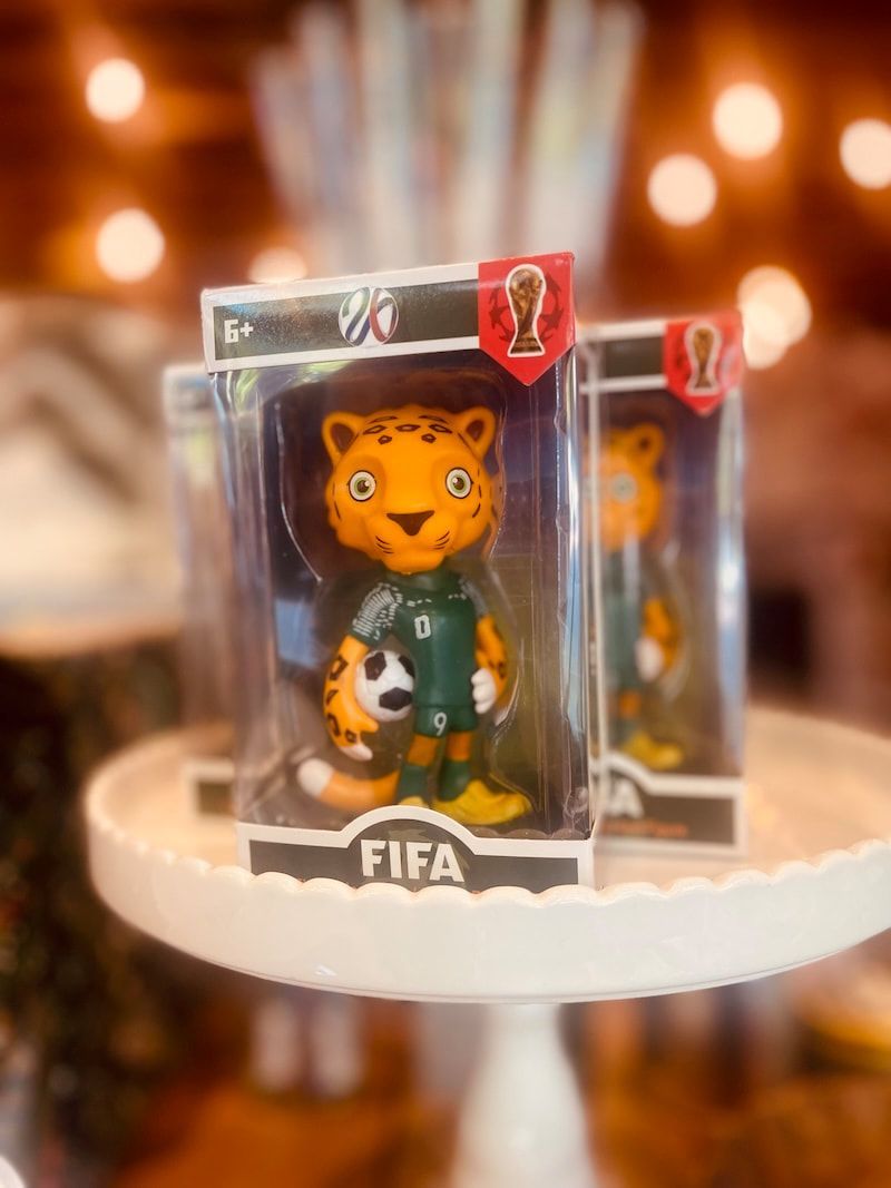 Small FIFA mascot figurines displayed in clear boxes on a round white shelf with warm lights behind.