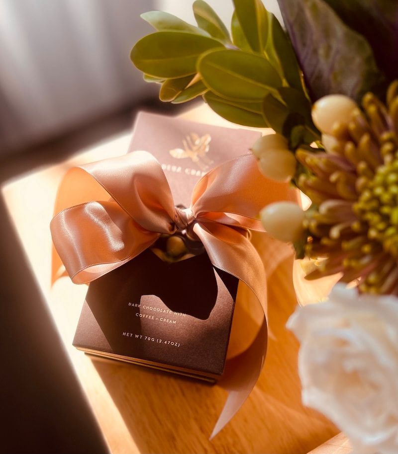 A dark gift box with a pink satin bow and green leaves on a sunlit wooden table.