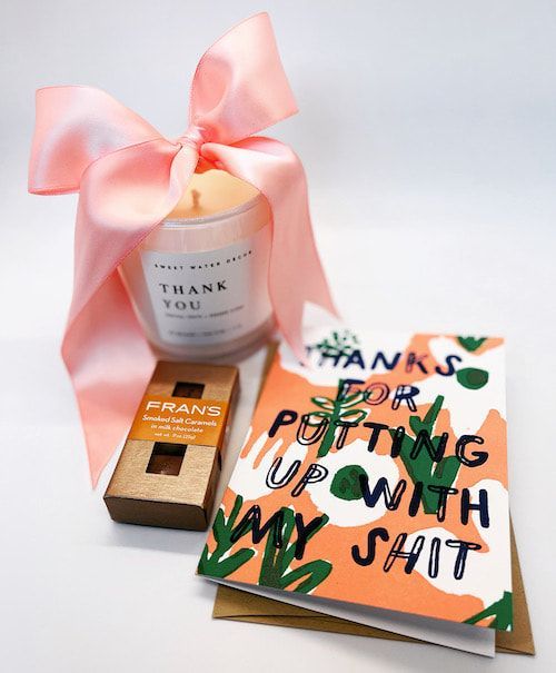 Gift set with pink ribbon, candle, chocolate, and card reading “Thanks for putting up with my shit.”