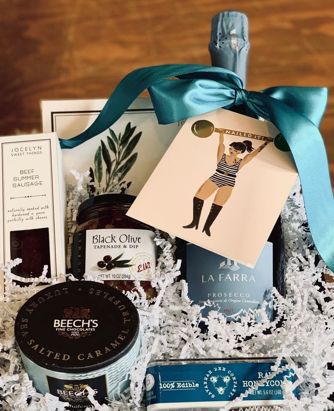 Gift basket with teal ribbon, olive oil, olives, soap, and packaged snacks in white crinkle paper.