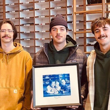 Three people indoors, one holding a framed blue-toned photo, with wooden shelves behind them.