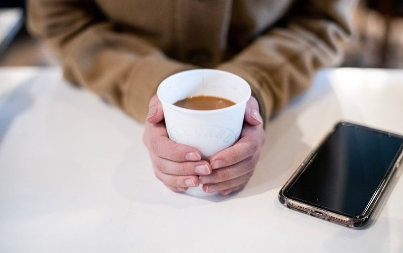 Hands holding a white coffee cup beside a smartphone on a white table
