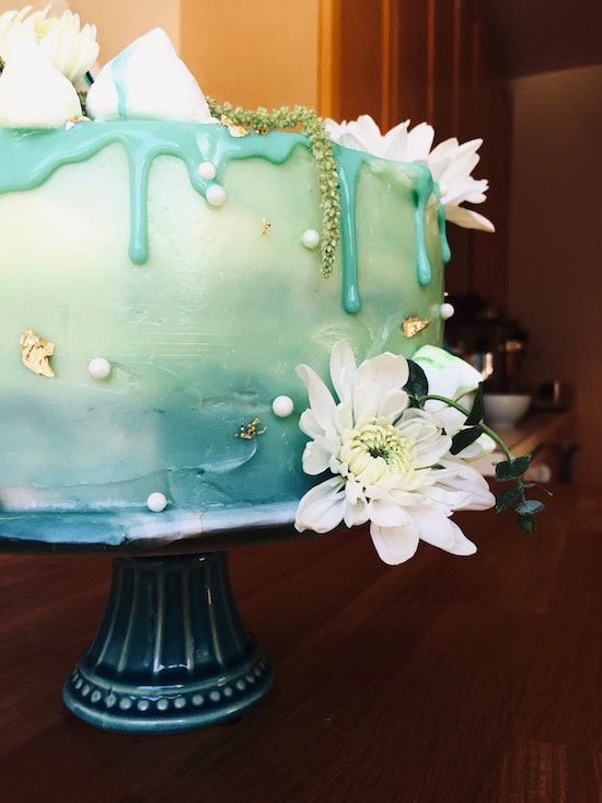 Turquoise frosted cake on a teal stand, decorated with white flowers and pearl-like accents