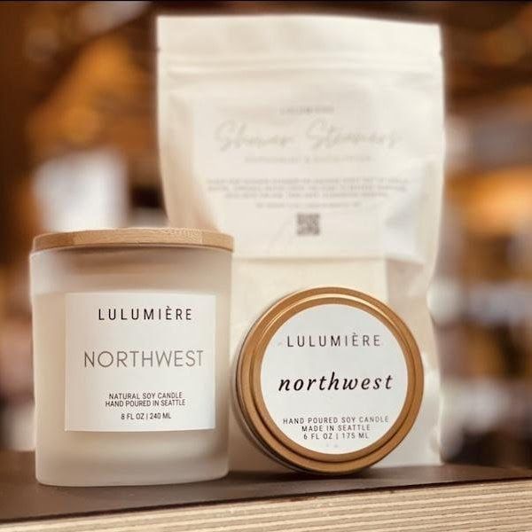 Two Lumière Northwest frosted candles with beige lids before a matching pouch on a warm blurred background