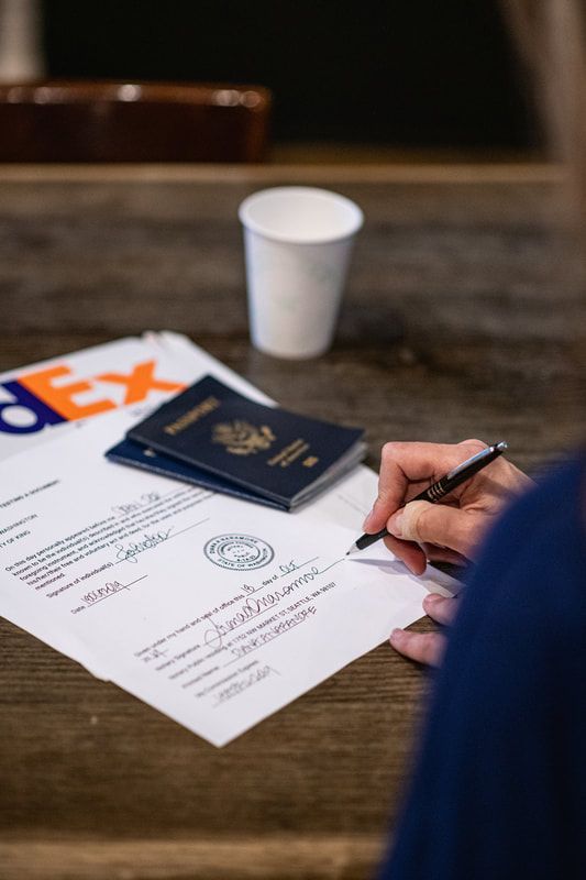 Person signing a document on a wooden table beside a passport, FedEx envelope, and coffee cup