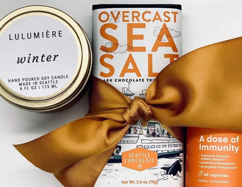 Assorted holiday self-care gifts with gold ribbon: winter candle, overcast sea salt chocolate, and immunity dose package