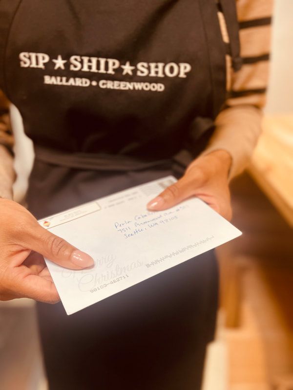 Hands holding a white envelope in front of a person wearing a Sip & Ship Shop apron.