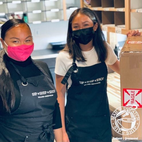 Two masked volunteers in aprons stand beside stacked boxes in a warehouse.