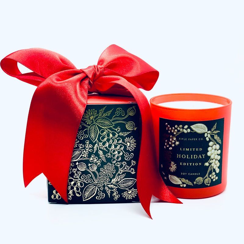 Red ribbon tied around a dark holiday gift box beside an orange scented candle on a white background