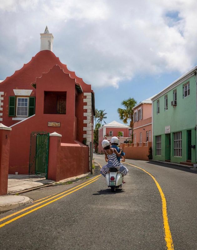 Scooter rider on a colorful street with red, green, and pink buildings under a cloudy sky