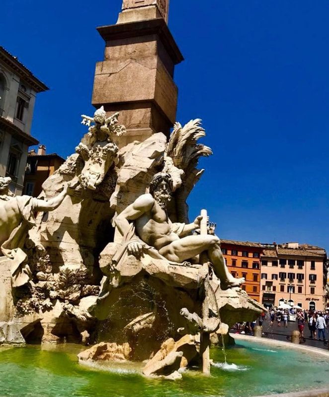 Stone fountain with reclining marble figures in a sunny plaza, blue sky and colorful buildings behind