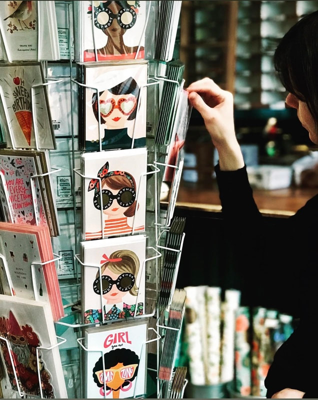 Postcard rack with illustrated faces in sunglasses; a person browses cards in a shop.
